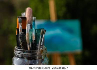 Many brushes in a glass jar with an easel with a painted canvas behind - Powered by Shutterstock