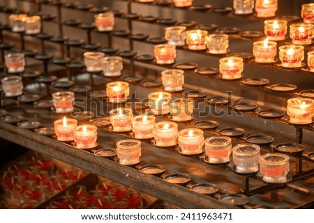 Many burning sacrificial candles or devotional candles in a church in small jars. The candles are placed on a special candle holder and most of them are lit.