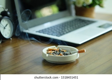 Many Cigarette In White Ashtray On The Desk With Laptop Computer. Smoking To Relieve Stress At Work But Unhealthy