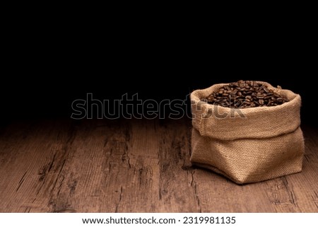 The many coffee beans and bag and scoop are placed around on a wooden table in a warm, light atmosphere, on dark background, with copy space.