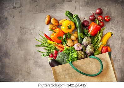 Many Colorful Vegetables Overflowing From Fallen Beige Grocery Bag Over Rustic Scratched Grey Background