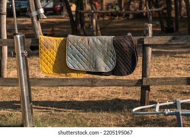 Many Different Saddle Pads Hanging On The Railing And Drying After Tournament. Removable Saddle Pads For Horse In Fresh Air. Horse Equipment Hanging On A Fence On Farm In Sunset.