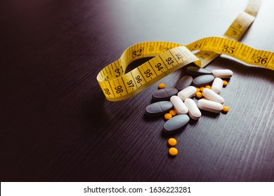 Many different weight loss pills and supplements as food with fork and spoon with measuring tape on black wooden table. Diet pills and supplements, prescription weight loss drugs. - Shutterstock ID 1636223281