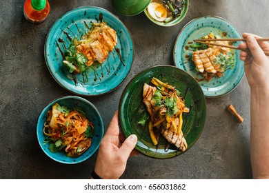 Many dishes of asian foods on dark table with human hands holding dish with asian seafood