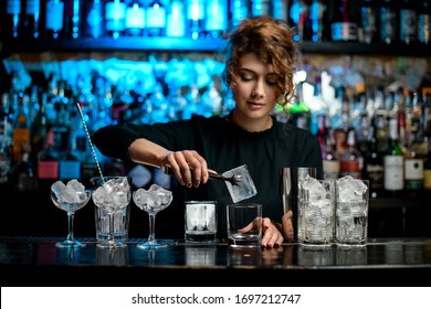 Many glasses with ice stand on bar counter. Bartender lady put large piece of ice with tongs to glass.