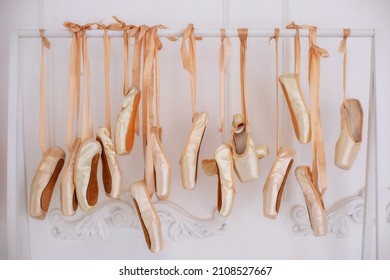 Many hanging ballet shoes on white wall background in studio. New pointe shoes with satin ribbons hanging on rank. Ballet shoes hang on bar in room. Concept of dance, ballet school, ballerinas clothes