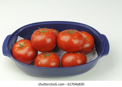 Many hydroponically grown red tomatoes grouped in an oblong blue bowl on a white quartz kitchen counter up close. Several red tomatoes with stems in a blue dish.