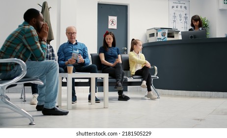 Many Patients Sitting In Waiting Area Lobby At Hospital Reception, Preparing To Start Checkup Visit With Appointment. Mother With Child, Senior Man And Other People In Waiting Room. Tripod Shot.