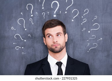 So Many Questions! Confused Young Man Standing Against Blackboard With Question Marks