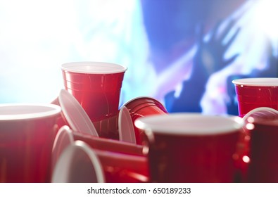 Many Red Party Cups With Blurred Celebrating People In The Background. College Alcohol Containers In Mixed Positions. Marketing And Promotion For Events Or Beer Pong Tournament.