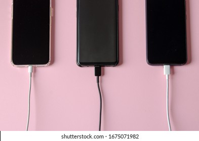 Many Smartphones Are Charging With White Usb Wires On Pink Table. Top View Of The Place For The Text. Flat Lay