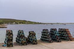 Many Stacks Of Cleaned Crab Traps And Lobster Pots On The Dock Of Blacksod Harbor With Boats Anchored In The Bay In The Background