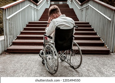Many stairs. Horizontal image of depressed disabled man sitting in the wheelchair and facing the difficulties with climbing the stairs alone
