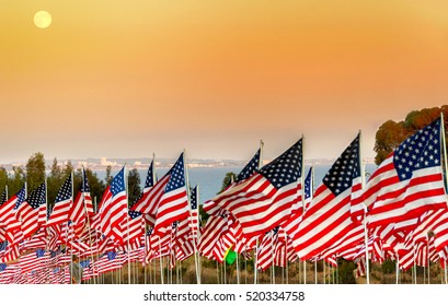 Many USA American flags on poles flying outdoors with ocean and coastline background and sunset sky.