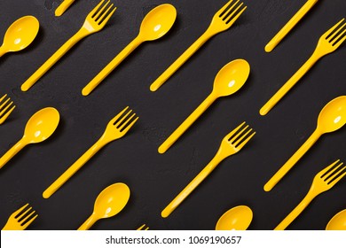 Many yellow plasic forks and spoons on black background, top view. Disposable table wear concept