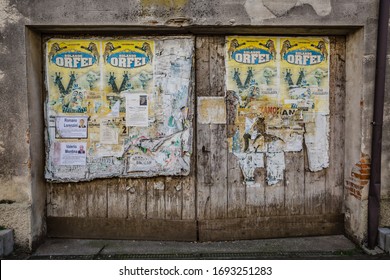 Manzano, Italy - 25.03.2020: Cityscape. View Of Old Wooden Gate Used As A Bulletin Board: Old Posters, New Funeral Notices