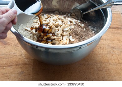 Maple syrup is being poured into a steel mixing bowl containing the raw ingredients for granola: oats, sesame seeds, sunflower seeds, ground flax seeds, and cashews. - Powered by Shutterstock