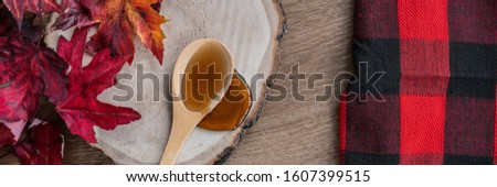 Maple syrup sweet ingredient for desssert recipes, liquid dripping from wooden spoon on wood log rustic sugar shack banner panoramic with buffalo plaid and red leaves.
