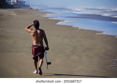 Mar Del Plata, Argentina, January 5, 2021. Young Adult Male Walking Along The Beach By The Sea With A Skateboard In Hand And Drinking Something. Boy With Cap On Head, No T-shirt And Tattoos.