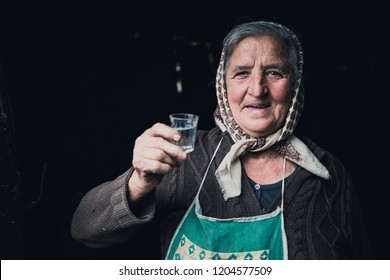 MARAMURES, ROMANIA - SEPTEMBER 10, 2018: Portrait Of An Old Eastern European Grandmother Lady With A Head Scarf With A Small Glass Of An Alcoholic Drink Who Looks In The Camera And Wants To Say Cheers