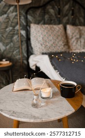 Marble coffee table with aromatic diffuser, open paper book, scented candle and cup of tea over bed in room. Cozy home atmosphere.