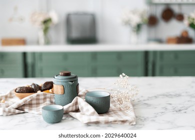 Marble Kitchen Island, Teal Teapot Set With Couple Small Cups Next To Bowl Filled With Dried Fruits And Nuts Laying On Tablecloth, Green Counter With Drawers And White Top In Blurred Background