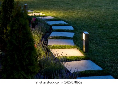marble path of square tiles illuminated by a lantern glowing with a warm light in a backyard garden with a flower bed and a lawn copy space.