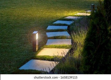 Marble Path Of Square Tiles Illuminated By A Lamp Glowing With A Warm Light In A Night Garden With A Flower Bed And A Lawn, Nobody.