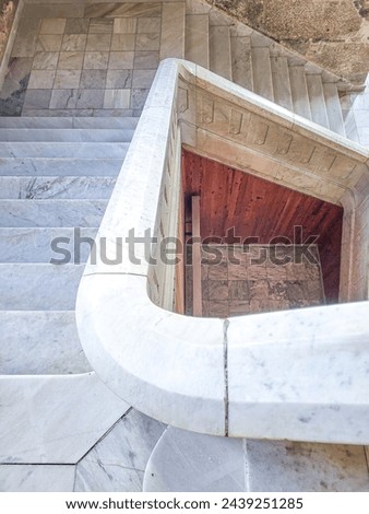 Marble stairs going down with handrails turning clockwise, wooden hall in the background and stone wall