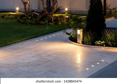 Marble Tile Playground In The Backyard Of Flowerbeds And Lawn With Ground Lantern And Lighting In The Warm Light At Dusk In The Evening.