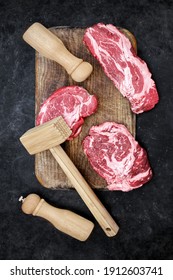 Marbled Raw Loin Beef Steaks, Wooden Hammer, Salt And Pepper Mill On Cutting Board. Beef Steaks BBQ Or Grilling, Overhead View. Raw Striploin Marbled Beef Steaks On Black Background, Top View.