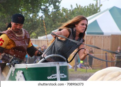 MARCH 18 2017 - RENAISSANCE FESTIVAL - DEERFIELD BEACH, FL  A Female Warrior Accompanied By A Male Warrior Leads Horses In A Carriage In A Sandy Pit In Front Of Festival Tents And Trees.