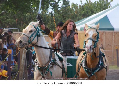 MARCH 18 2017 - RENAISSANCE FESTIVAL - DEERFIELD BEACH, FL  A Female Warrior Leads Horses In A Carriage In A Sandy Pit In Front Of Spectators, Festival Tents And Trees.