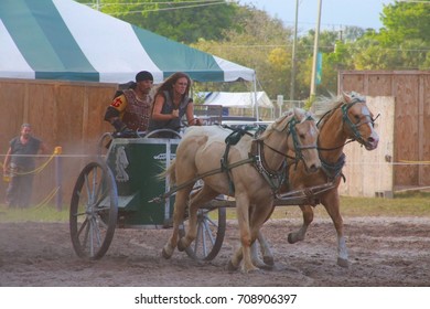 MARCH 18 2017 - RENAISSANCE FESTIVAL - DEERFIELD BEACH, FL  A Female Warrior Leads Horses In A Carriage In A Sandy Pit In Front Of Festival Tents And Trees.