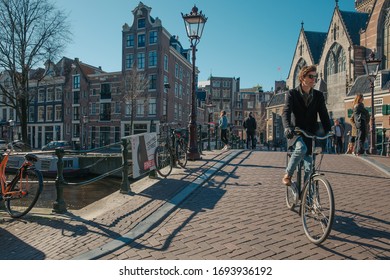 March 2020 - Amsterdam, The Netherlands - a young man is cycling over one of the beautiful De Wallen district bridges with old church in the background.