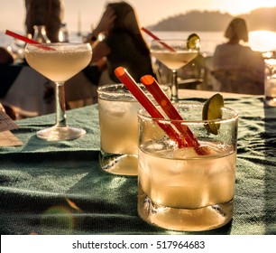 Margarita Cocktail Glasses On A Beach Bar Table With People, Sunset, Sea And Mountains Background