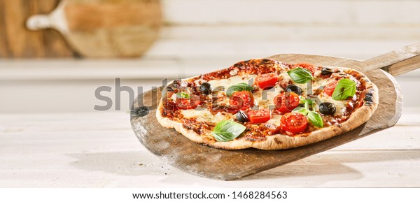 Margherita Italian pizza with\
extra trimmings topped with fresh tomato, olives and shaved\
parmesan cheese served on an old wooden pizzeria paddle in panorama\
banner format