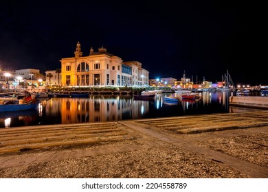 Margherita Theater And Fishing Row Boat In Old Harbor Of Bari, Italy, At Night. Colors And Lights Reflected On The Sea, Long Exposure.