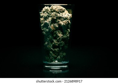 Marijuana buds in a clear glass cup with a black background a pile of high quality marijuana buds with some cigars of weed ready to smoke. Top view with background of black stone.