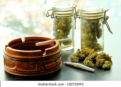 Marijuana Joints and Jars of Weed. A marijuana joint in an ashtray, with another joint, a pile and two jars full of more marijuana.