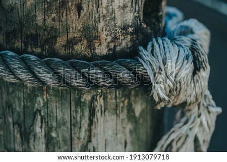 Marina distressed wooden pillar for tying up boats at a dock. Sailor's post and anchor setting. Maritime navy or pleasure boating use. Retro rope on old wooden stump close up. Boho artistic shot.