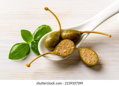 Marinated Caper Fruits In A Spoon And Basil Leaf On A White Wooden Background. Whole And Halved Pickled Caper Berry Close-up. Mediterranean Cuisine Ingtedient. Sour Condiment And Garnish. Top View.