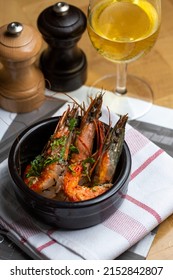 Marinated Fried Grilled Prawns With Fresh Herbs In Black Plate On Wooden Table With Rustic Kitchen Towel And Fresh Newspaper, With Glass Of White Wine And Salt Pepper On The Background