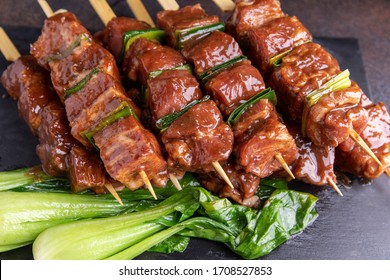 
Marinated pork and leek skewers on wooden skewers in a brown marinade, semi-finished product, on a dark board with Bok-cho