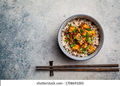Marinated tofu with sesame seeds, green onions and brown rice in a bowl on a gray background. Asian food concept.