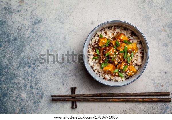 Marinated tofu with\
sesame seeds, green onions and brown rice in a bowl on a gray\
background. Asian food\
concept.