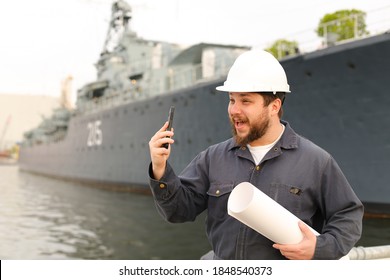Marine assistant engineer holding VHF walkie talkie and blueprints near vessel in background, wearing helmet and work jumpsuit. Concept of maritime profession , job and seaman.