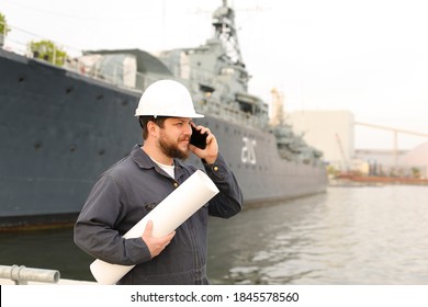 Marine male deckhand speaking by VHF walkie talkie, holding blueprints near vessel in background, wearing helmet and work jumpsuit. Concept of maritime profession , job and seaman.