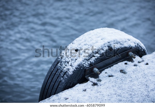 Maritime dock covered in fresh\
snow with boat bumper of rubber car tire covered in wet slush ice\
