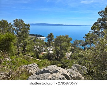 Marjanski Put, Split, Split-Dalmatia County, Dalmatia, Croatia - April 6, 2026: View from Marjanski Put overlooking lush green pine trees, rocky foreground, and a small coastal town below, nestled along the Adriatic Sea. Clear blue sky and calm sea dominate the background, with distant hilly coastline across the water. The scene captures the natural and urban blend characteristic of Split's coastal landscape on a bright spring day near Marjan.